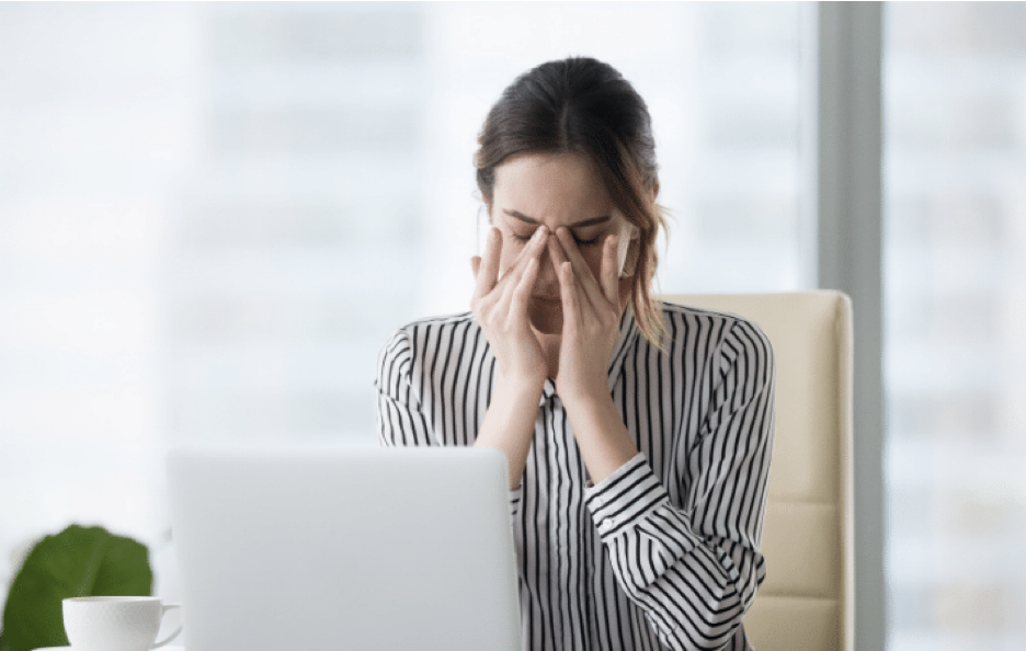 A woman sitting in front of a laptop computer, that is rubbing her eyes and the ridge of her nose in pain.