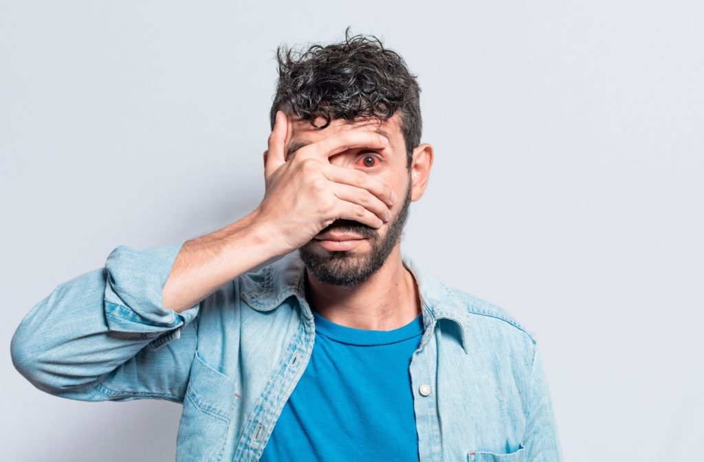 A person covers their right eye with their right hand, while their left eye is open, showing a red and irritated ocular surface.