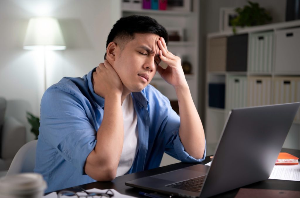 A person sitting in front of a laptop experiencing a headache and neck pain.