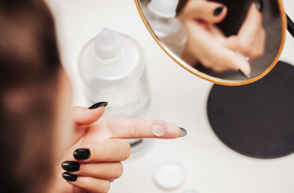 A person holds a contact lens on the tip of their finger in front of a small mirror with a bottle of solution nearby.