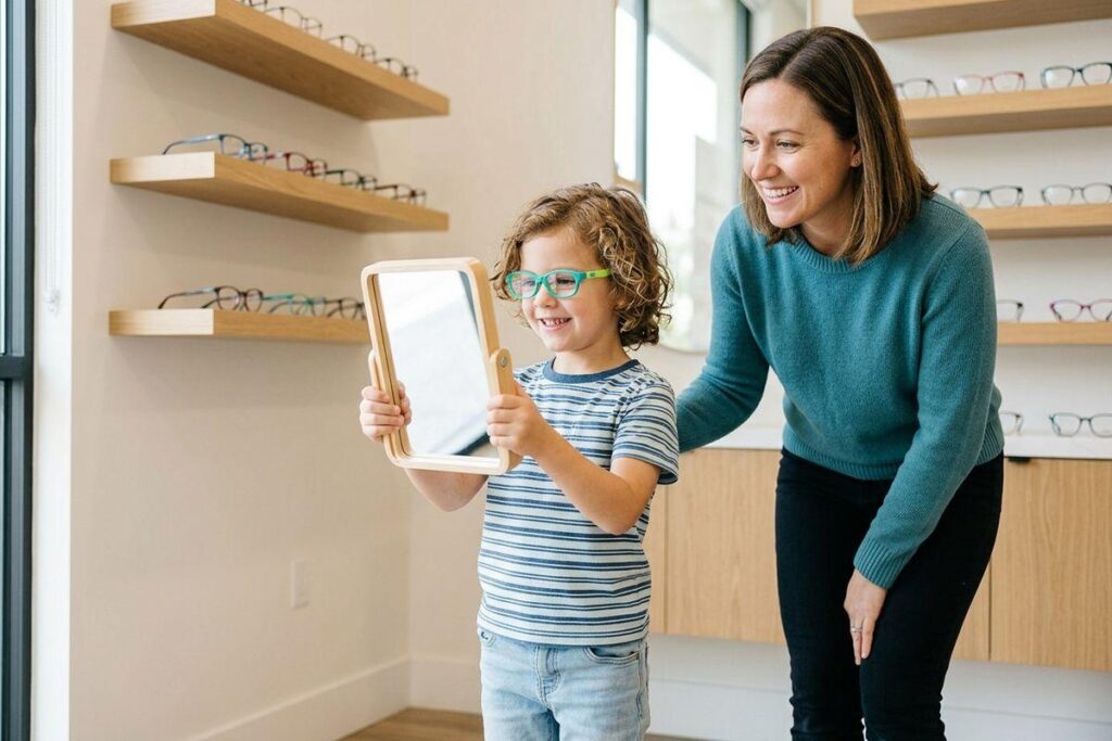 Child wearing green glasses holds a mirror, smiling, in an eyewear store while an adult looks on smiling, shelves of frames behind them.